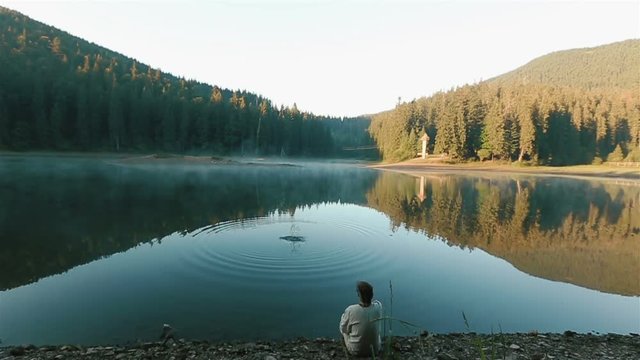 Handsome young thoughtful man in embroidery shirt throwing stones into the water and looking on the picturesque mountain lake Synevir. Ukrainian mythology concept. Back view