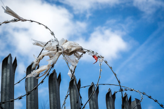 Steel fence with barbed wire with rags, plastic and cloth against blue sky.
