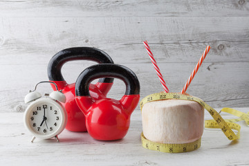 Two red kettlebells with measuring tape, drinking coconut, straws, and vintage clock on rustic white wooden table. Healthy diet and fitness concept.