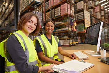 Two female colleagues in a warehouse office look to camera