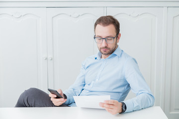 Fototapeta premium Businessman sitting at his desk, looking at paper document and holding smart phone 