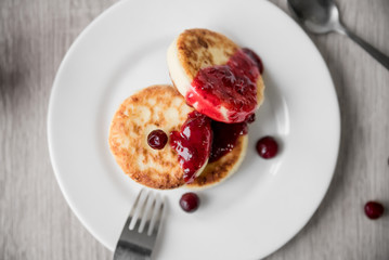Cheese pancake with cranberry sauce on a white plate on a wooden table, top view