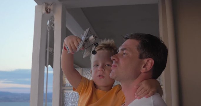 Little boy boy playing with toy plane while father holding him in arms. They standing on the balcony with view to the sea. Leisure with dad