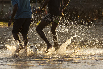 Legs of black children playing footbal in water at sunset