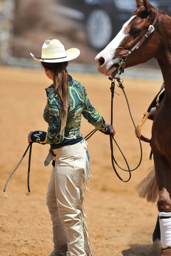 The western style rider with cowboy chaps and hat leads her horse from the field