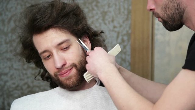Close up man's face at the barber's - the barber is cutting man's beard with the scissors and the comb