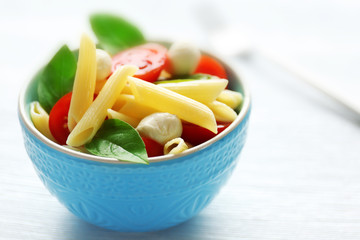 Cooked penne pasta with mozzarella, fresh tomatoes and basil in blue bowl on wooden table
