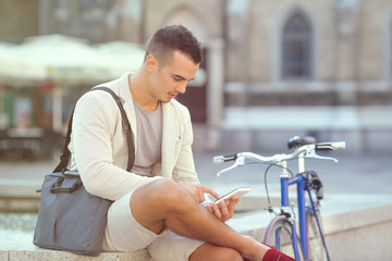 Smiling young businessman sitting on the street next to his bicycle and using digital tablet