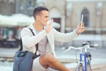 Handsome young businessman is relaxing in the city and waving to camera of his digital tablet. He is sending photo messaging or having a video call.