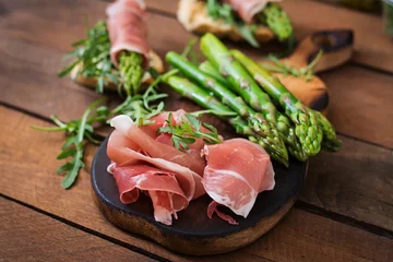 Fototapeten Parma ham, asparagus and arugula on a wooden background © timolina