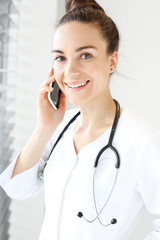 Atrakcyjna lekarka rozmawia przez telefon. Lekarka ubrana w biały kitel rozmawia przez telefon stojąc przy oknie. - obrazy, fototapety, plakaty