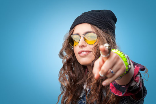 Closeup portrait of beautiful trendy hipster girl with long curly hair pointing finger at you and smiling wearing checkered shirt, black beanie hat and glasses on blue background.Youth style,fashion.