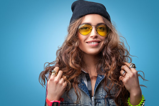 Closeup portrait of beautiful trendy hipster girl with long curly hair and hands up smiling wearing  checkered shirt, denim vest, black beanie hat and glasses on blue background.Youth style,fashion.