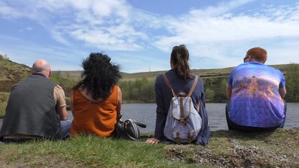 Family of four people or two couples, or group of four adults sitting relaxing on a lake shore and enjoying sunny day out