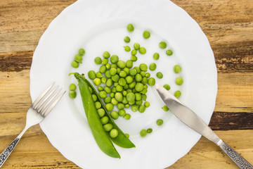 Fresh green peas on plate with fork and knife closeup, on wooden surface