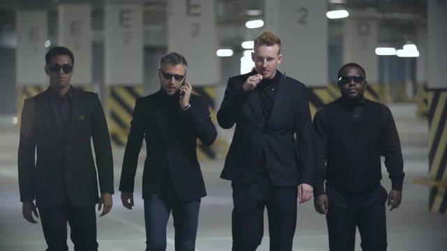 Four brutal men in black suits and sunglasses walking towards the camera in slow motion; one of them talking on the phone