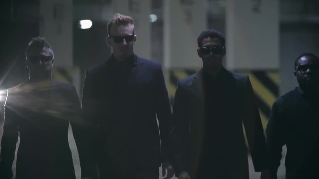 Four gangsters in black suits and sunglasses walking towards the camera in slow motion