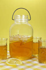An old fashioned mason jar containing iced tea. Three glasses have tea in as well. A yellow checked table cloth with lemon slices in the foreground.