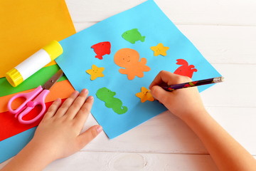 Child holds a pencil and draws. Child doing a card with sea animals and fish. Sheets of colored paper, scissors, glue, set for kids art