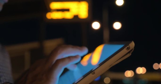 Close-up shot of man using tablet PC on bus station at night, led display with information in background