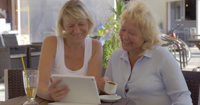 Two happy senior female friends sitting in outdoor cafe, having drinks and looking at photos on tablet computer