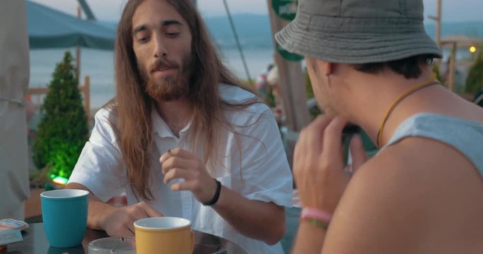 Two friends having a vivid talk in street cafe in the evening, long-haired man smoking and they clanging cups