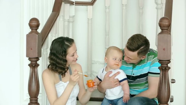 The mother feeds the child, and dad is holding him. Happy family. Mom dad and 6 month old baby. Family sitting on the stairs at home.