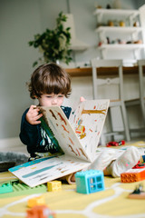 Little boy holds a book and sees the drawings. The child sits on the floor. Near scattered toys. The concept of family