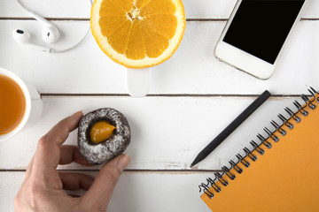 Hand holding chocolate cookie, white wood table top, carrot juice, earphones, slice of orange, smart phone, orange notebook, black pencil around

