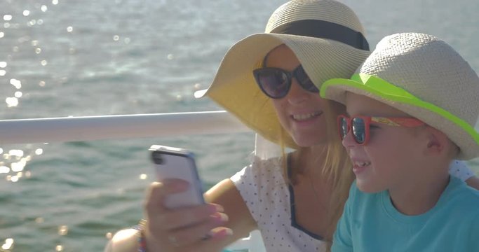 Happy young mother and little son in hats and sunglasses taking selfie on cell while traveling by sea. Then they looking through the photos