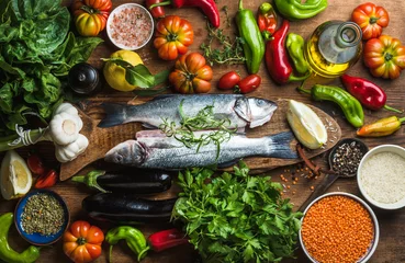 Foto op Plexiglas Rauwe ongekookte zeebaarsvis met groenten, granen, kruiden en specerijen op snijplank over rustieke houten achtergrond © sonyakamoz