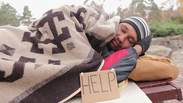 Sad homeless man suffering from cold, sleeping on bench covered with blanket