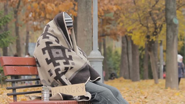 Lonely poor person sitting on bench, warming up with old blanket, man starving