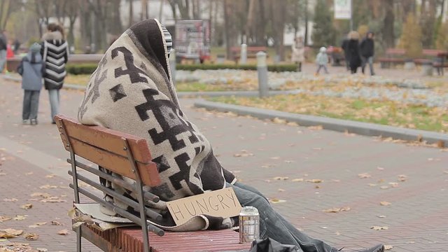 Hungry beggar sitting alone in cold park, many people passing by indifferently