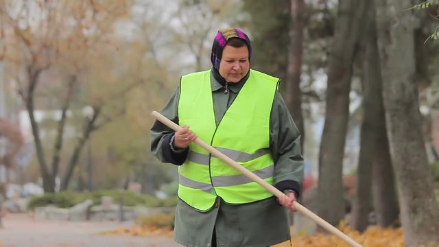 Unhappy overweight woman sweeping city park in autumn, hard-working person