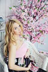 Young beautiful pregnant woman with long blond hair sitting in a wicker chair on the background of a decorative tree with pink flowers, look into the camera.