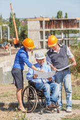 Disabled person in the construction helmet with documents in hand talking on the phone on the background of building. Successful wheelchair user directs the work.