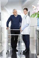 Fototapeta na wymiar Doctor Standing With Senior Man Using Walker In Rehab Center