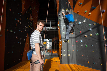 Fototapeta na wymiar Man stands on the ground near rock wall indoors, looking at the camera and insuring the climber, using rope and belay device