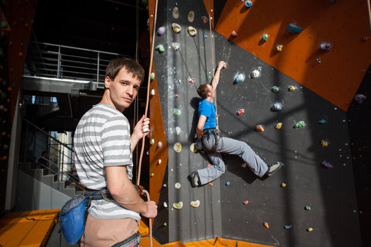 Man stands on the ground near rock wall indoors, looking at the camera and insuring the climber