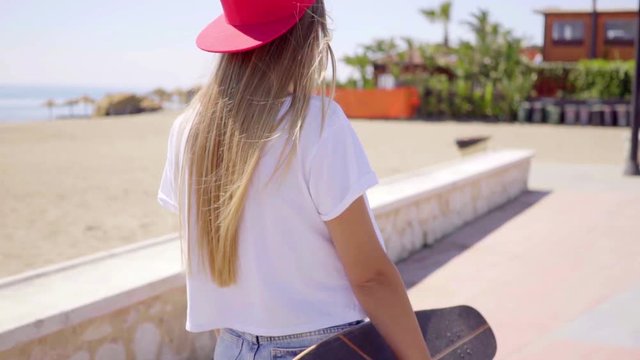 Rear view on single tough young blond female skateboarder in white shirt and backward red hat walking on sidewalk