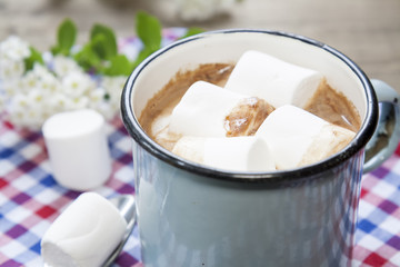 Hot cocoa chokolate with marshmallows in blue cup on checkered napkin and spring blossoms.