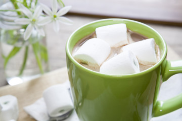 Hot cocoa chocolate with marshmallows in green cup on napkin and fresh spring white flowers on wooden table