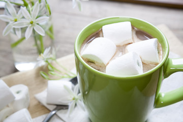 Hot cocoa chocolate with marshmallows in green cup on napkin and fresh spring white flowers on wooden table