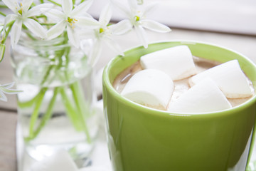Hot cocoa chocolate with marshmallows in green cup on napkin and fresh spring white flowers on wooden table