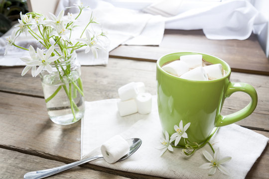 Hot cocoa chocolate with marshmallows in green cup on napkin and fresh spring white flowers on wooden table