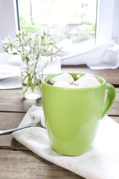 Hot cocoa chocolate with marshmallows in green cup on napkin and fresh spring white flowers on wooden table