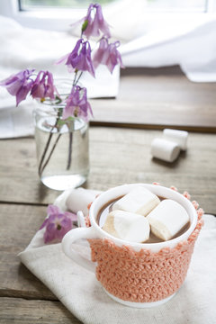 Hot cocoa with marshmallows in cup and cup crochet holder and fresh spring pink bell flowers on wooden table
