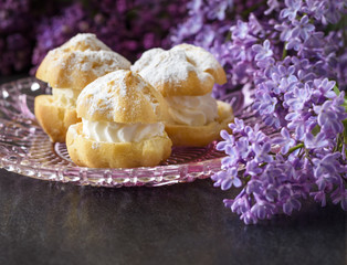 Cream puff or profiterole with filling, powdered sugar topping and lilac flowers isolated, on dark background