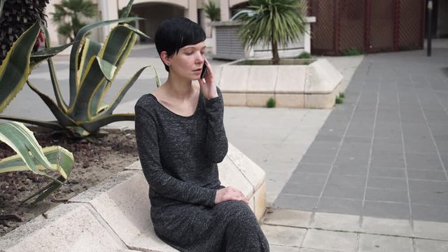 Women using mobile phone by her walking people. Female wearing in maxi grey dress. Caucasian model with black short hair talk on the phone in the open air.  